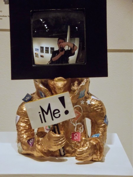 Photograph of a bust holding a sign that says "iMe!" with a mirror for a head. In the mirror is a photograph of Glen Farrelly taking a selfy.